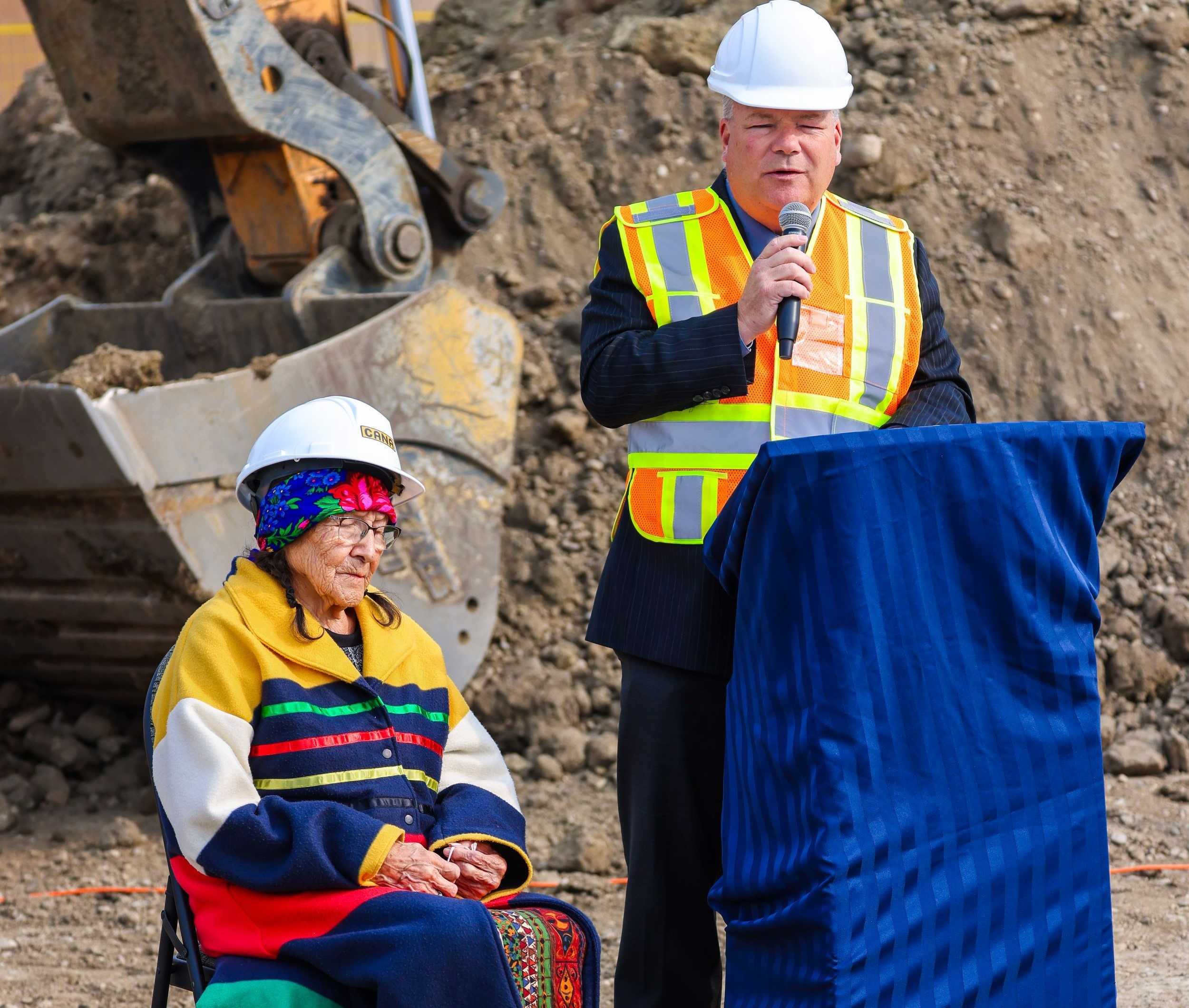 Mayor speaking at podium next to Blackfoot elder in foreground with large construction equipment in background